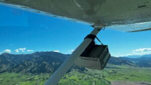 Aerial view from beneath a small aircraft flying over a green mountain valley, with part of the wing, support strut, and mounted equipment visible in the foreground.
