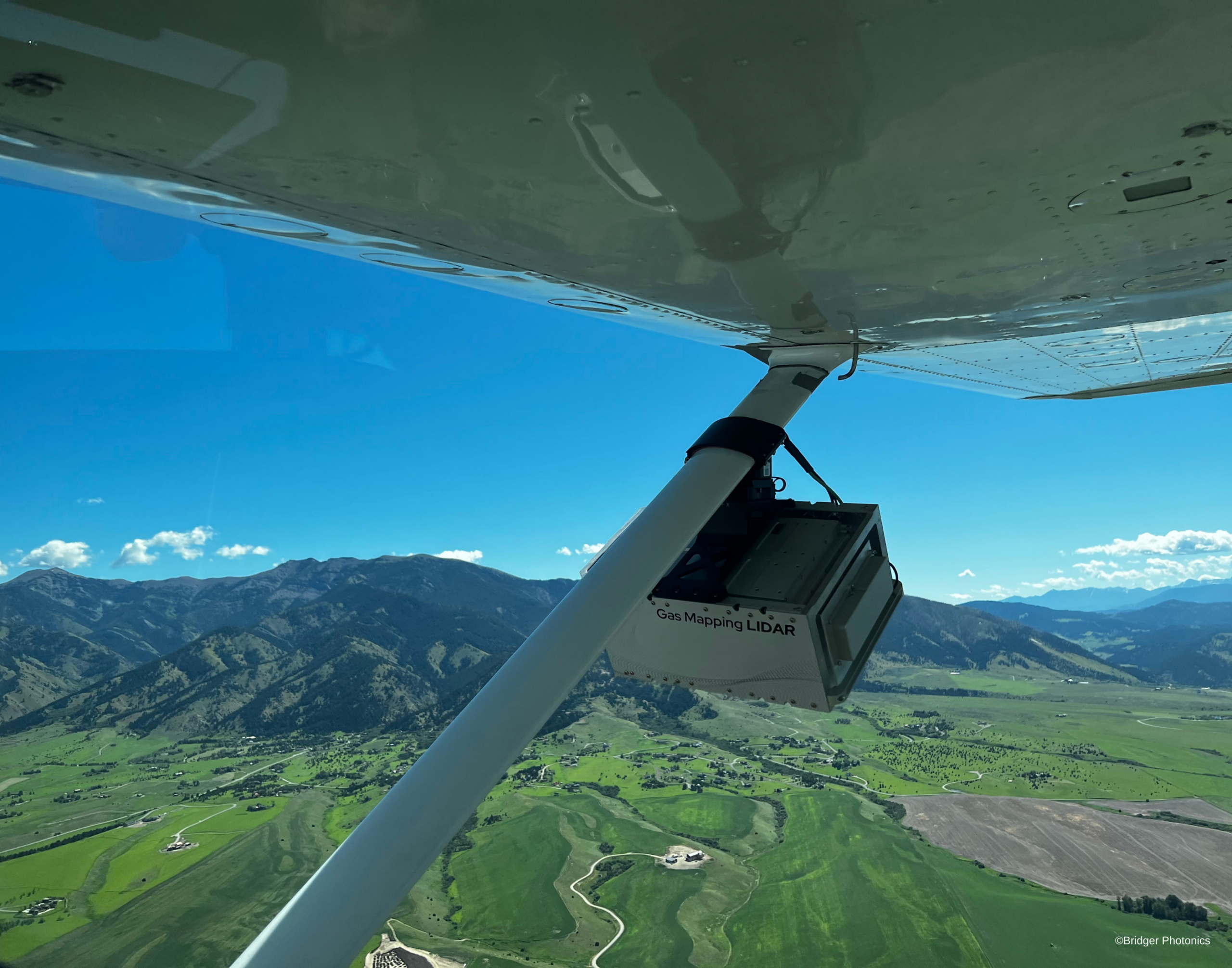 Aerial view from beneath a small aircraft flying over a green mountain valley, with part of the wing, support strut, and mounted equipment visible in the foreground.