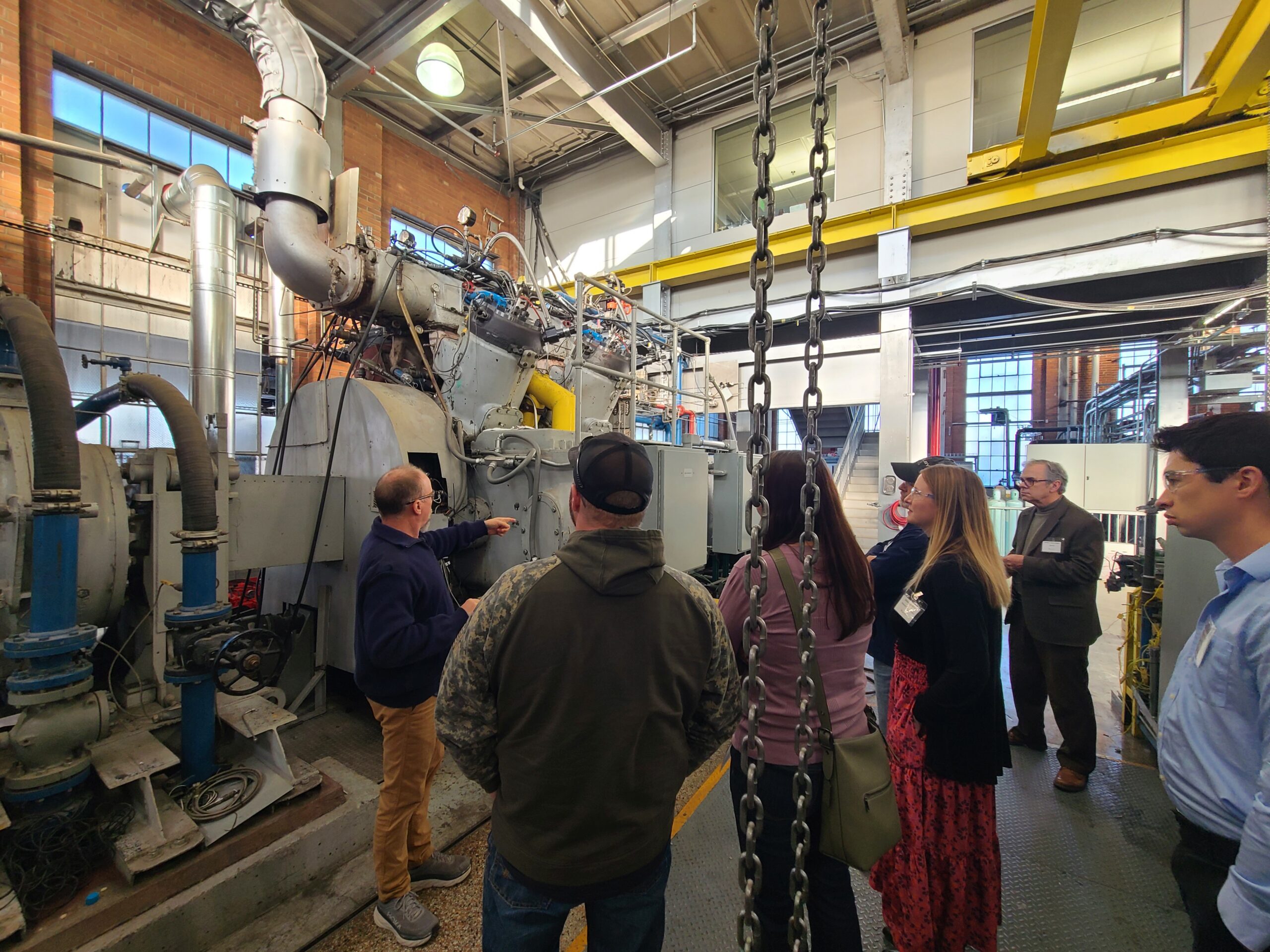 A group of people gathers around large industrial machinery while one person points out equipment components during a facility tour.