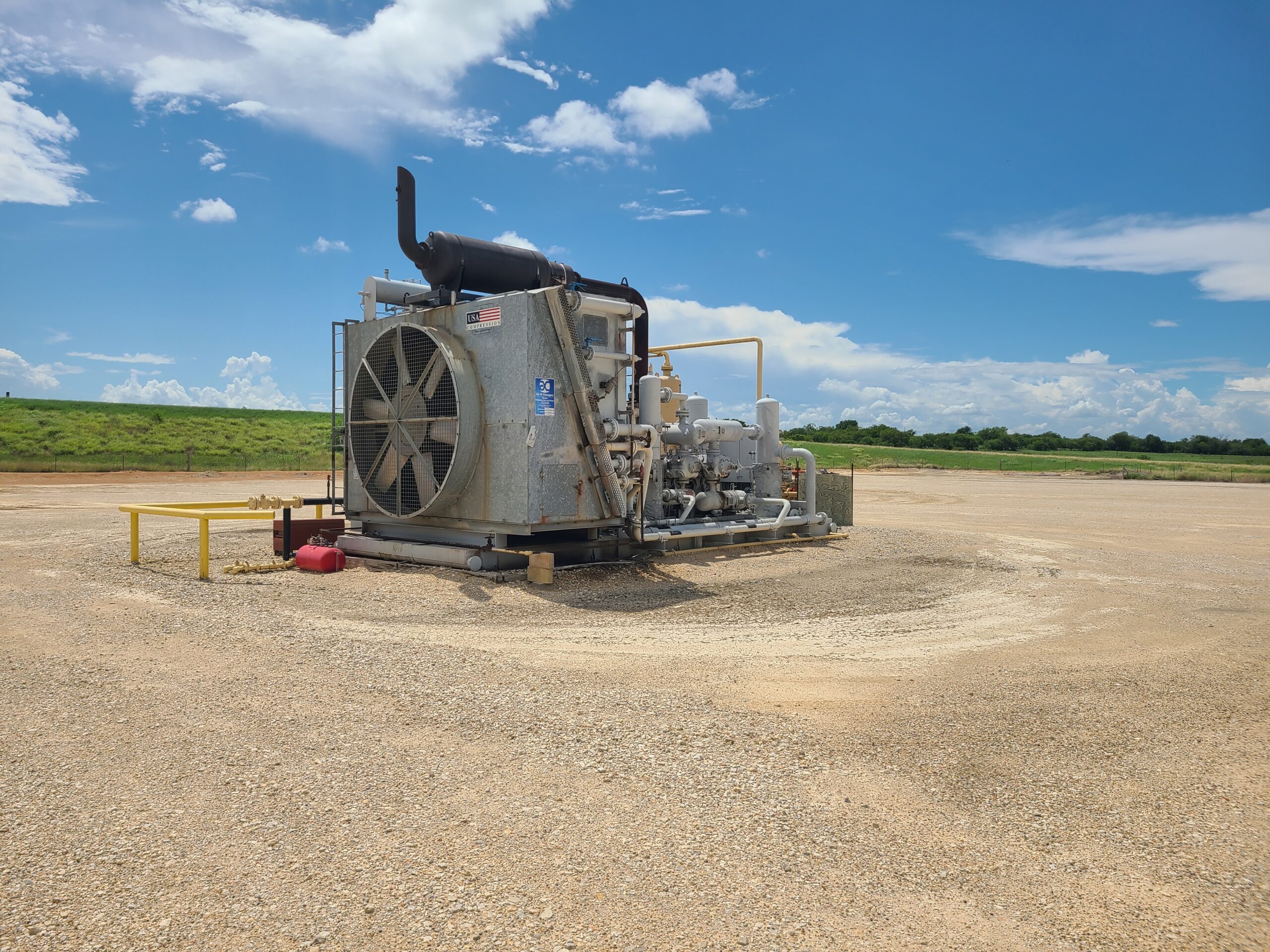Industrial compressor equipment and piping installed on a gravel pad beneath a blue, partly cloudy sky.