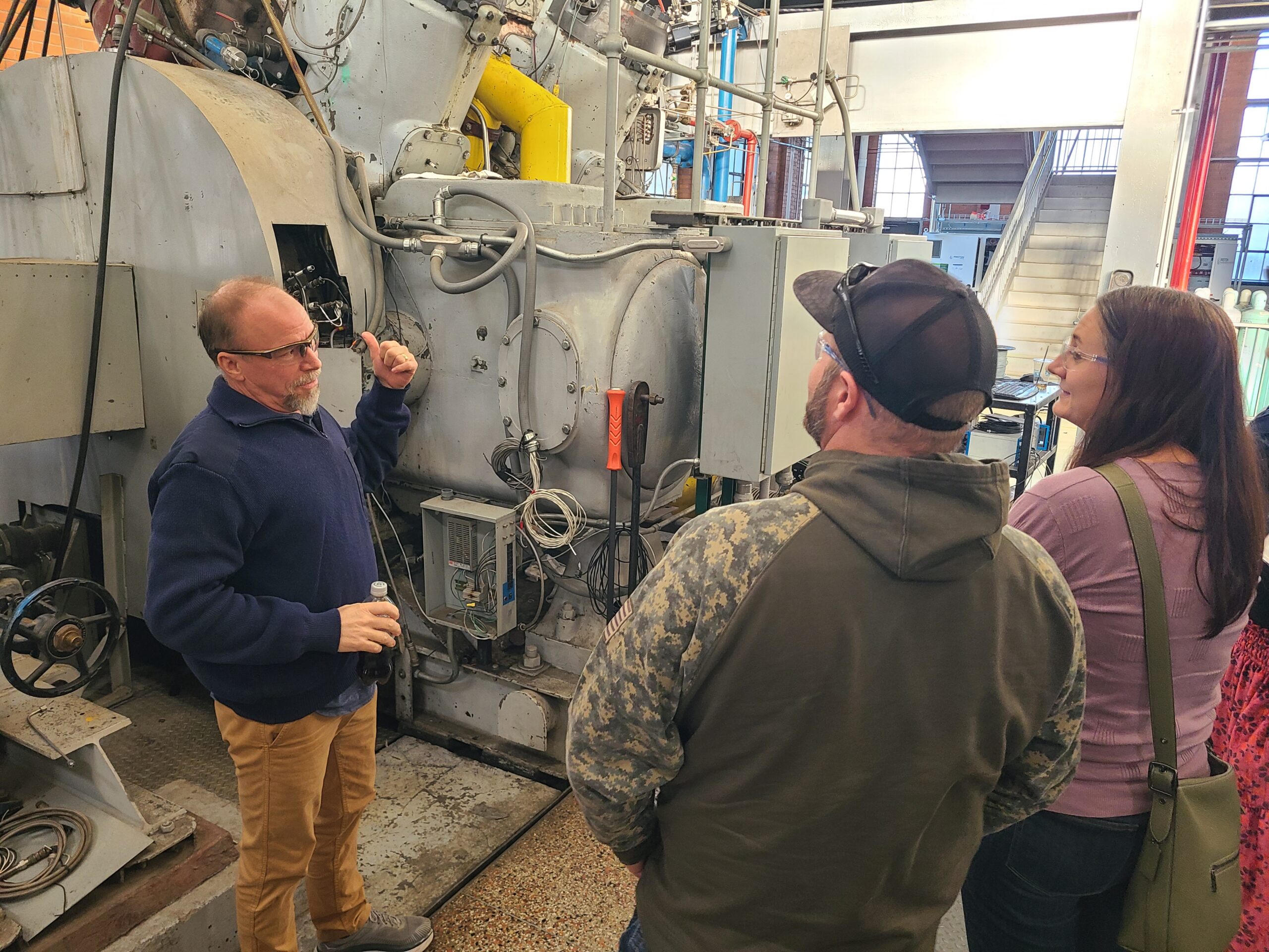 Three people stand beside a large industrial machine as one person gestures toward its equipment and components during a facility tour.