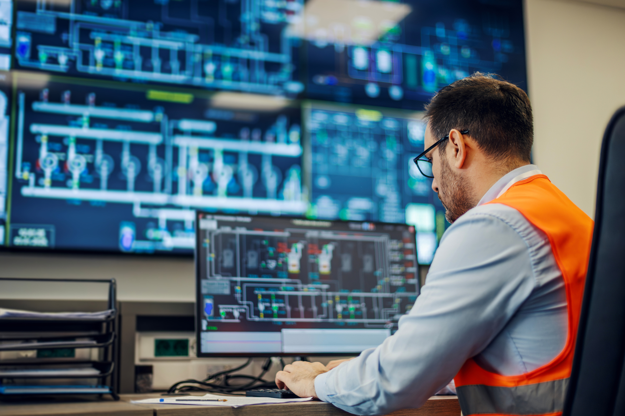 Male engineer wearing safety vest and glasses, sitting at a desk and operating a computer with multiple screens displaying complex industrial diagrams and data in a control room